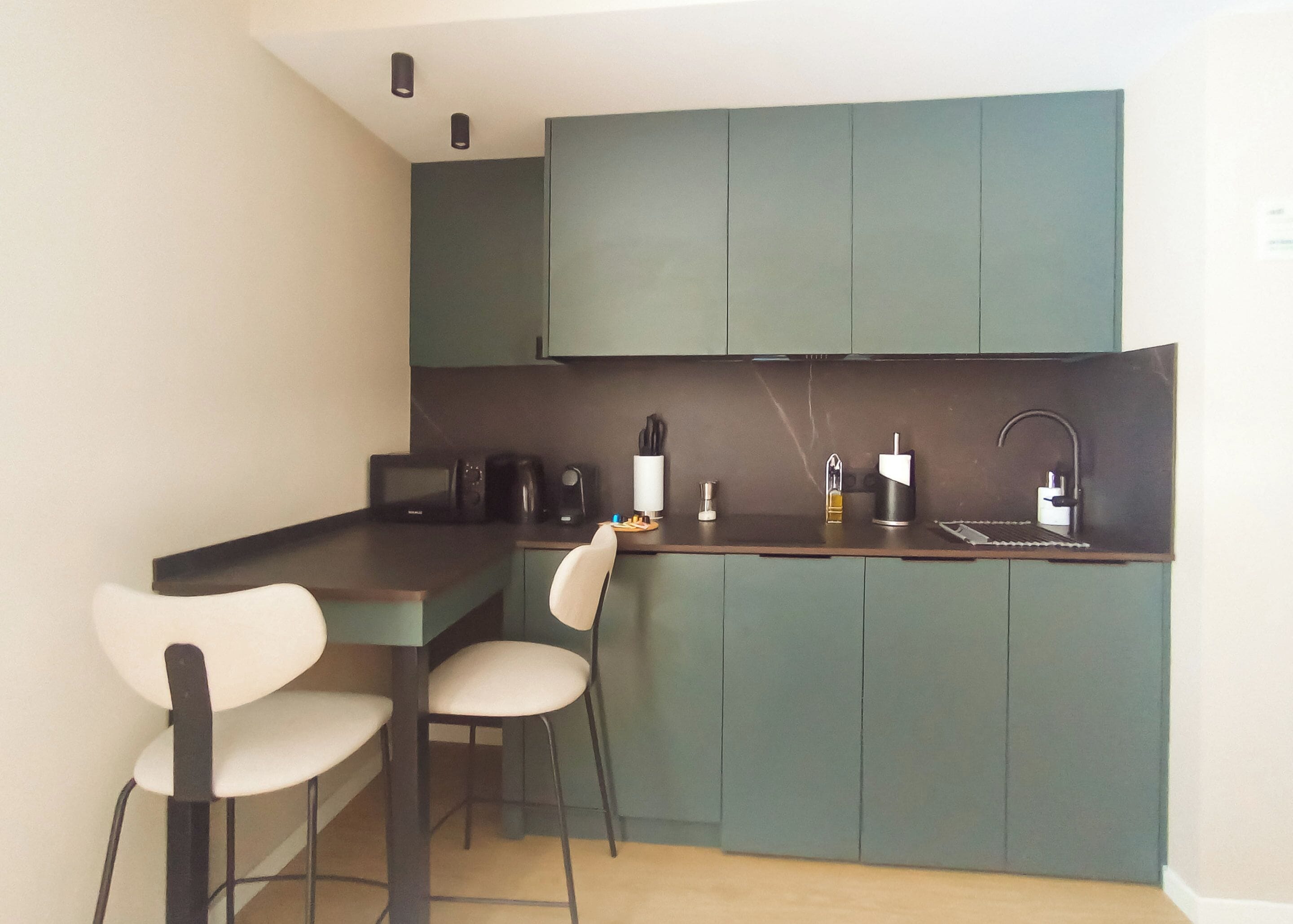 Una cocina compacta con gabinetes verde oscuro, una encimera negra y dos sillas blancas.