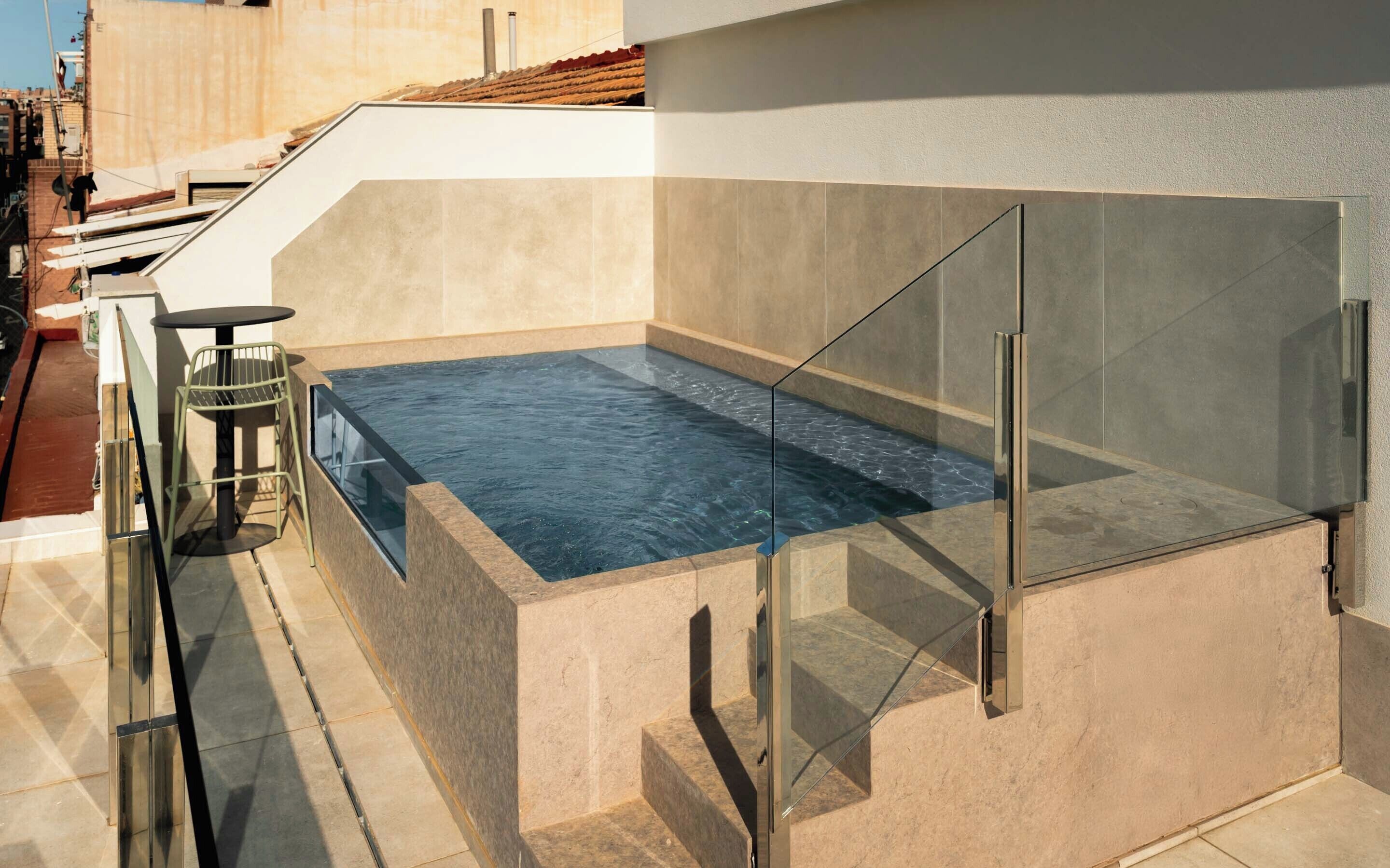 A rooftop swimming pool with glass railings, a small table, and chairs.