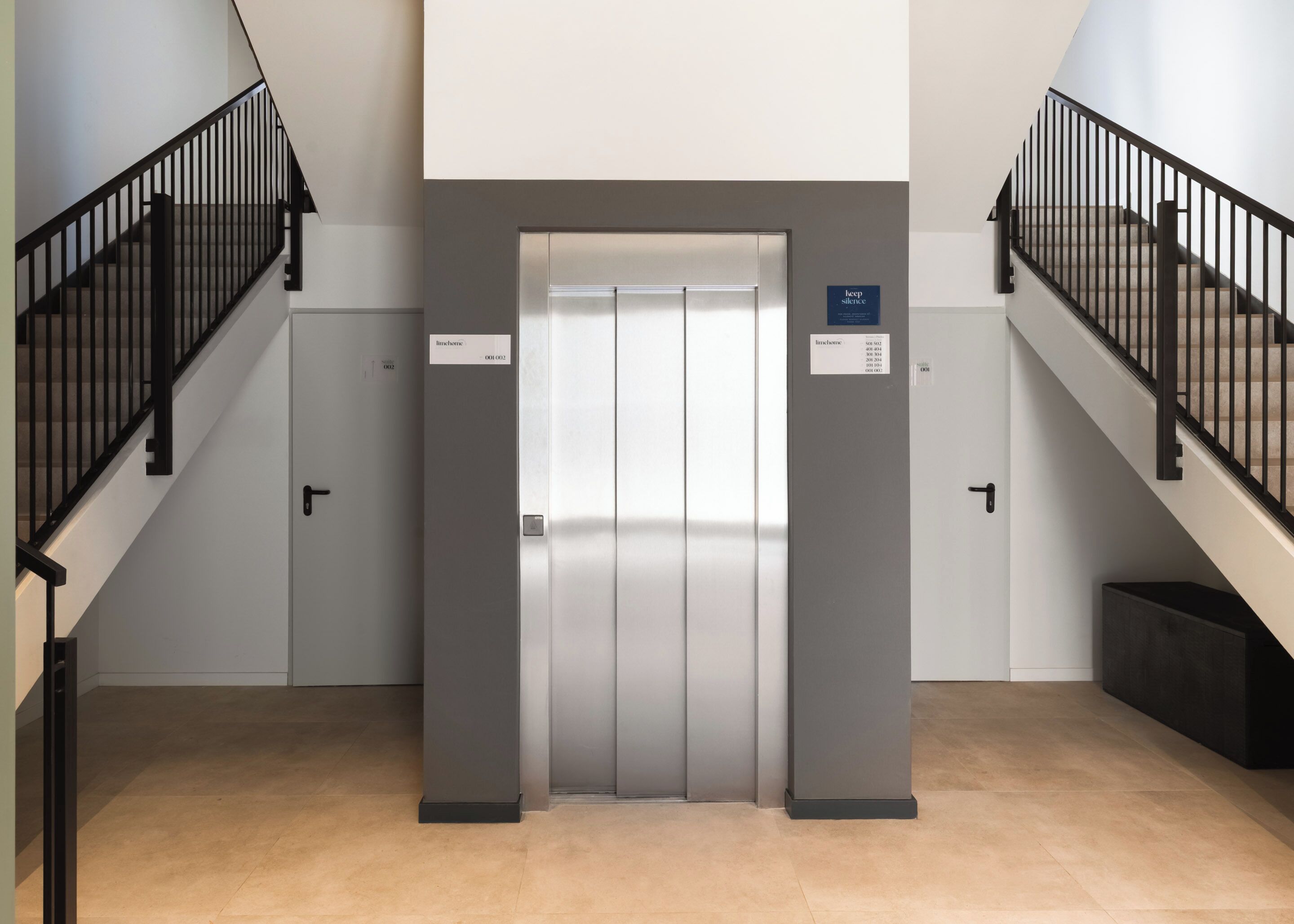 Een moderne lift met een metalen afwerking, omringd door zwarte wenteltrappen met metalen leuningen, en een vloer betegeld met beige tegels.