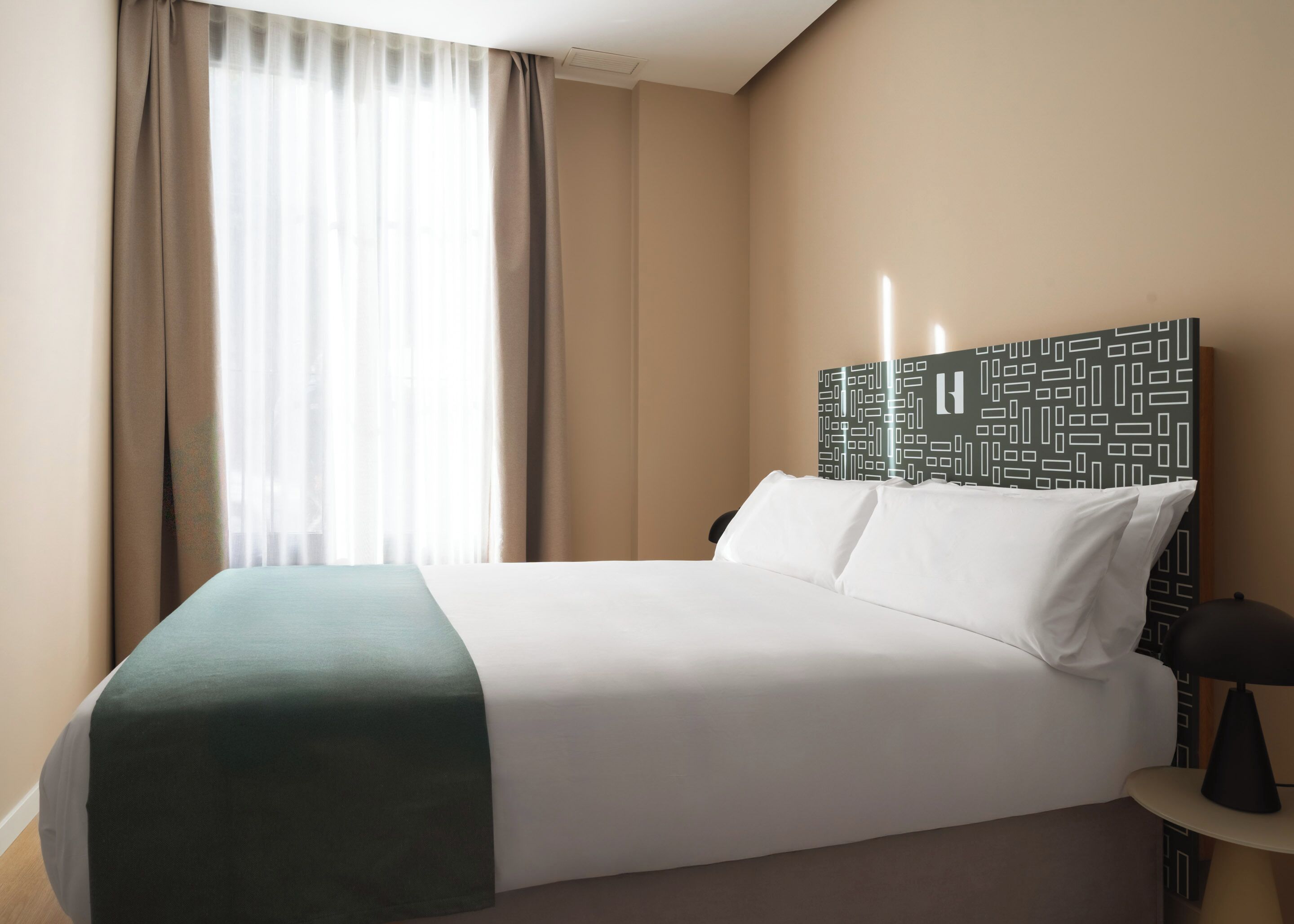 A hotel room with a bed, a bedside table with a lamp, a window with curtains, and a patterned headboard.