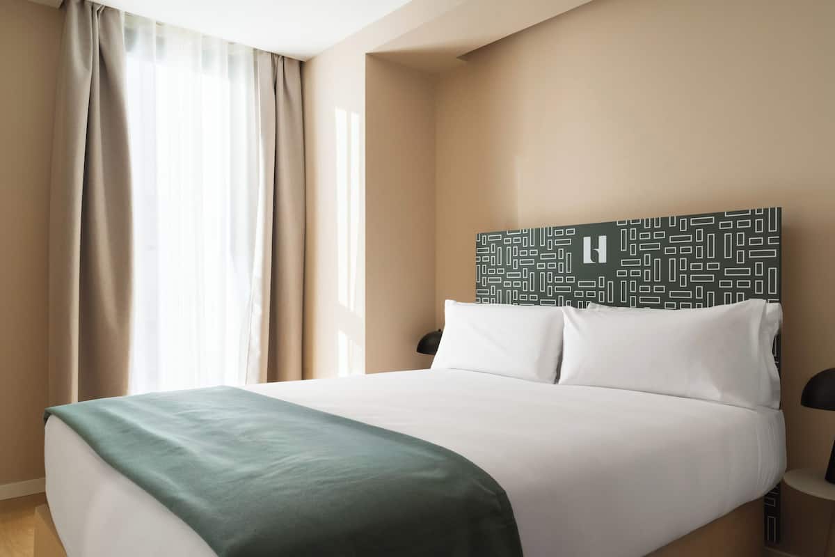 A hotel room with a bed, a headboard with a geometric pattern, a bedside table with a lamp, and a window with curtains.