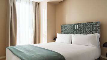 Une chambre d’hôtel avec un lit, une tête de lit ornée d’un motif géométrique, une table de chevet avec une lampe et une fenêtre avec des rideaux.