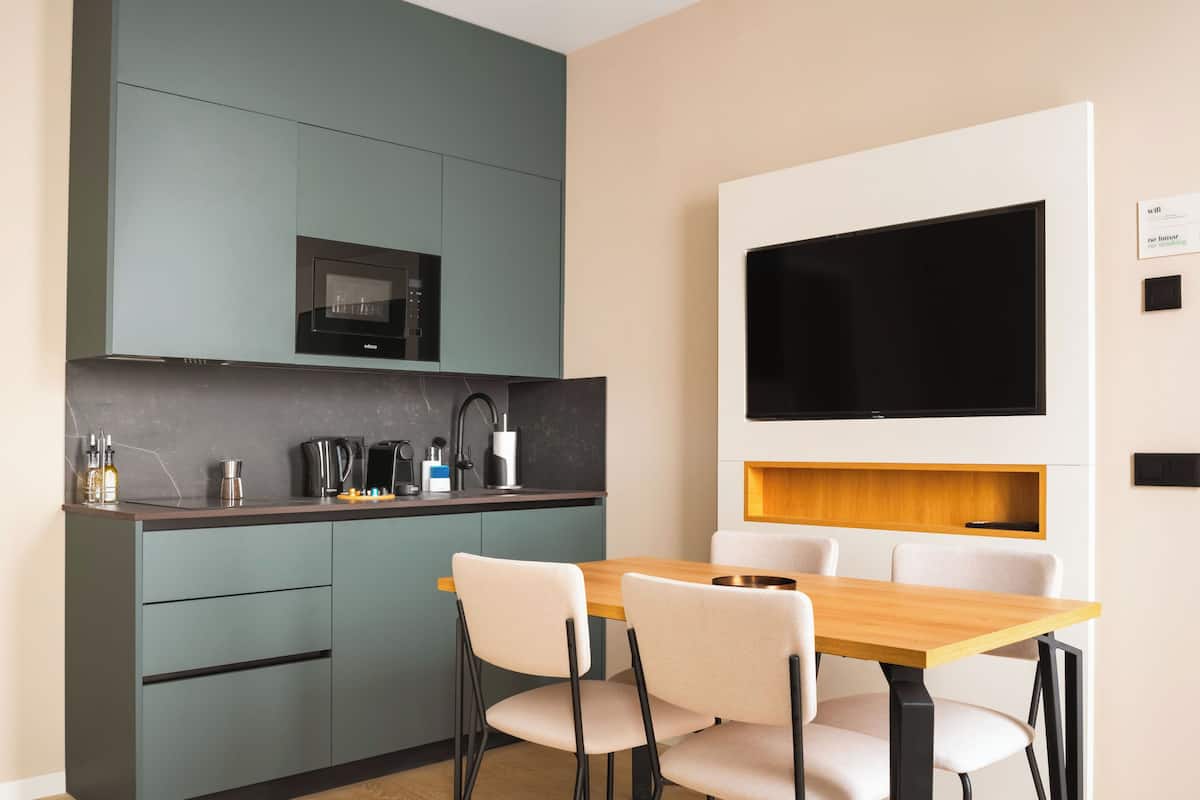 A modern kitchen with a built-in microwave, a coffee machine, and a kettle. There is a dining table with four chairs and a flat-screen TV mounted on the wall.