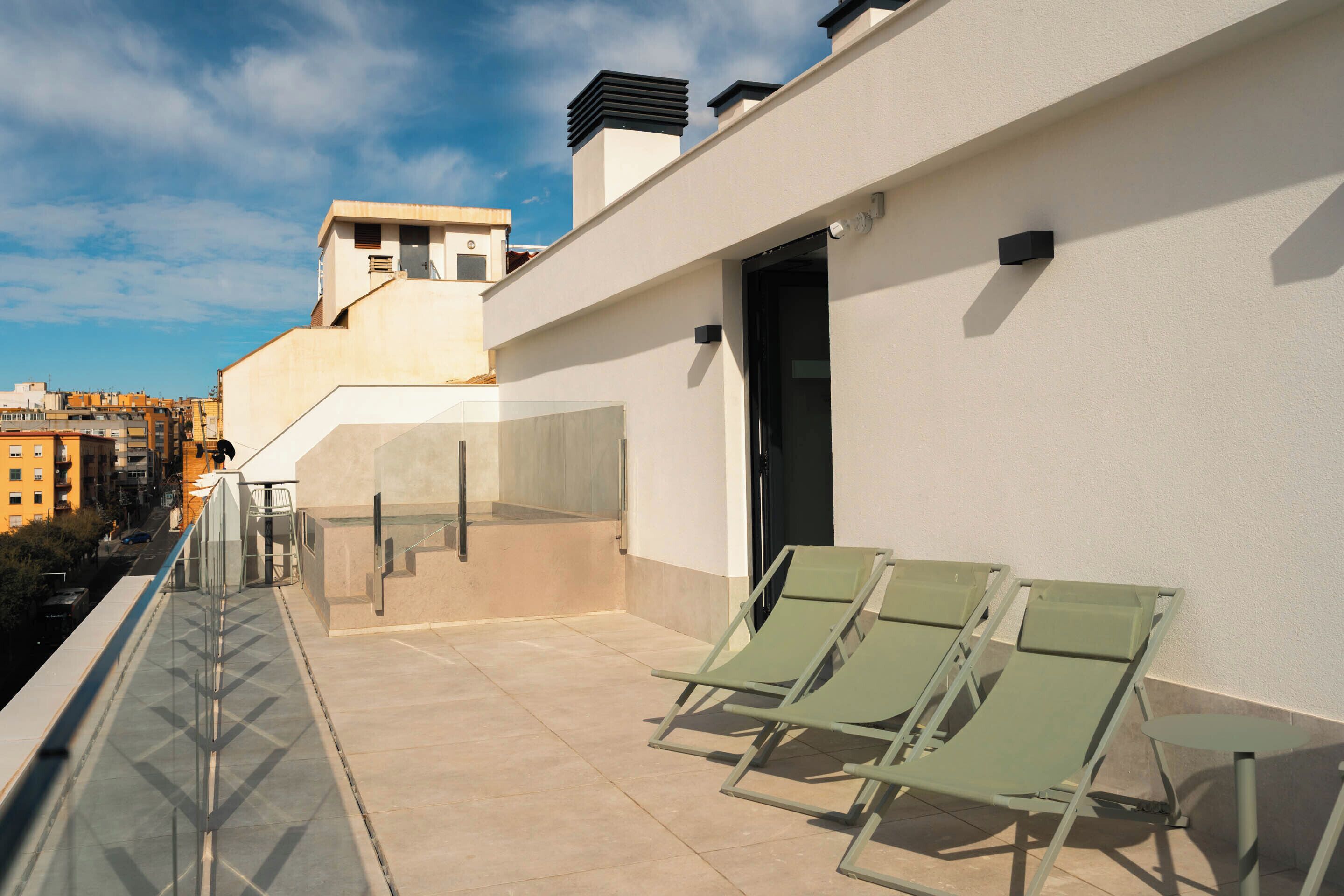 A rooftop terrace with a glass balustrade, two green lounge chairs, and a small table.