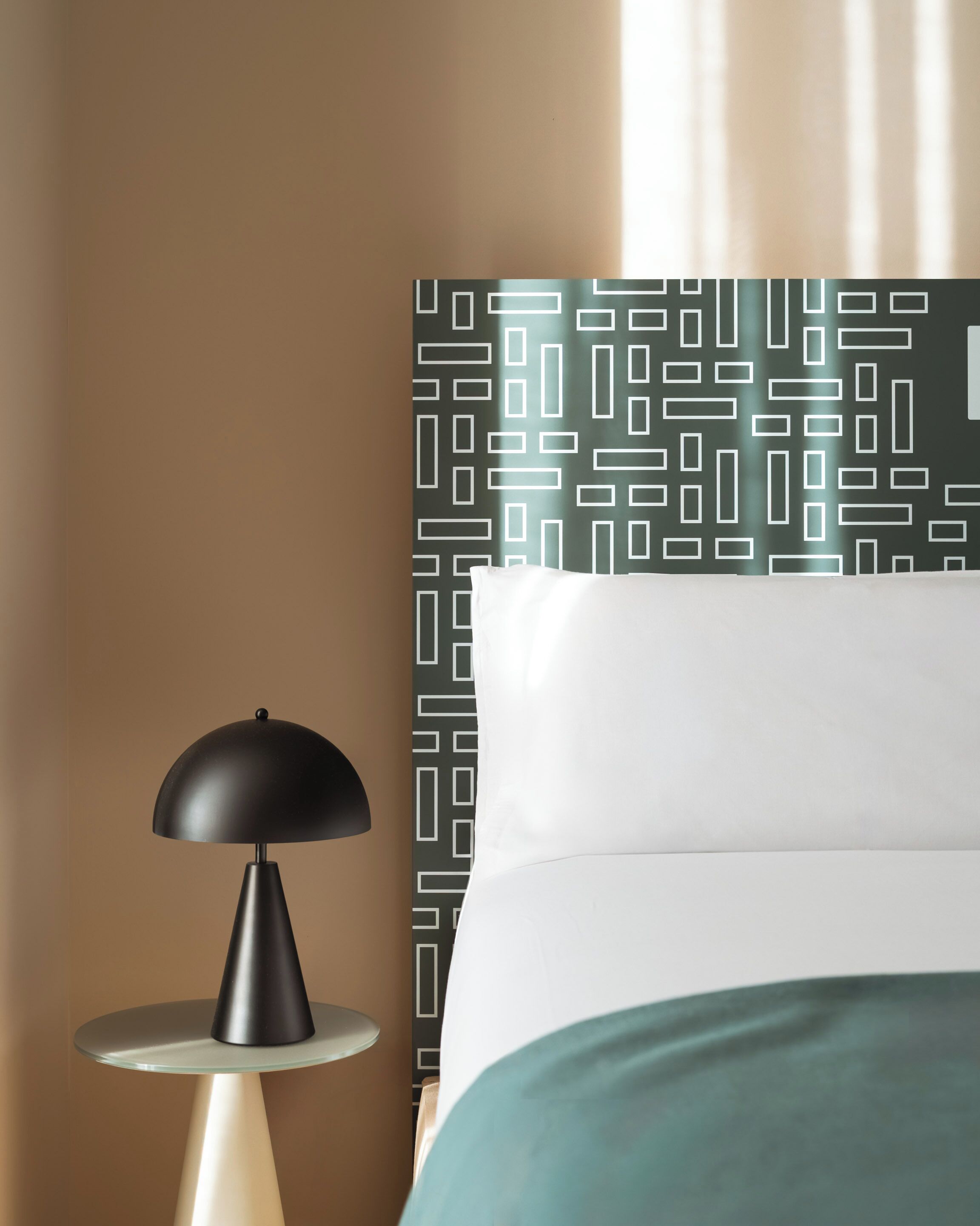 A neatly made bed with a green bedspread, a black lamp on a round table, and a patterned wall in the background.