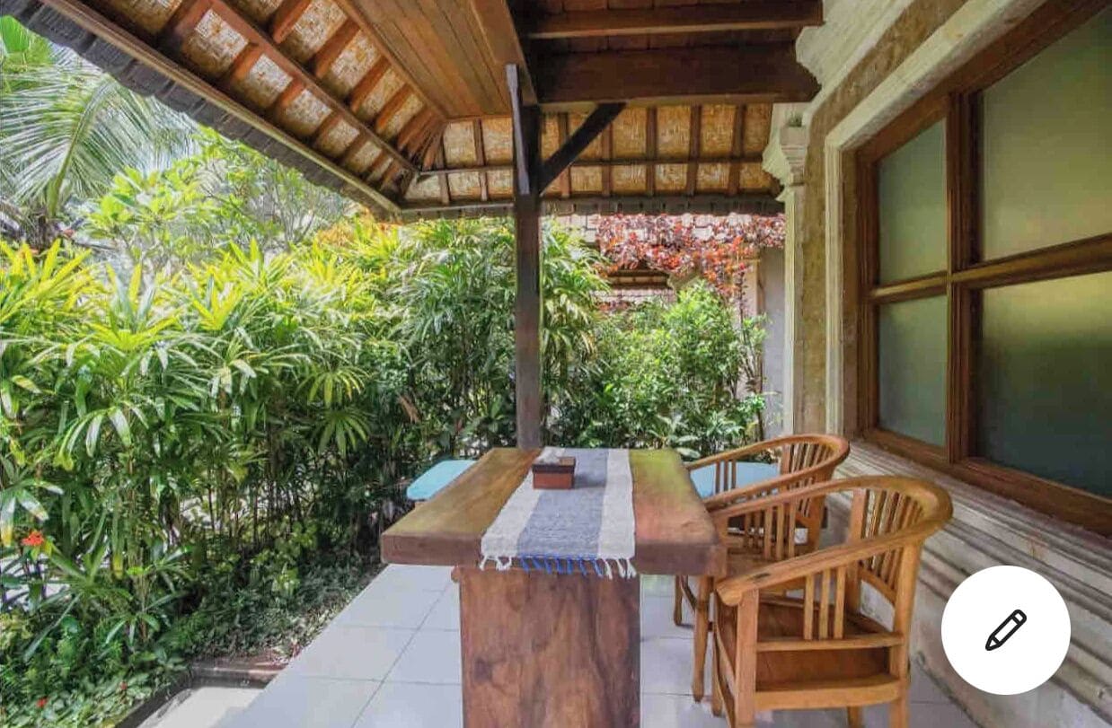 An outdoor patio with a wooden table and chairs, surrounded by lush greenery and a tiled floor.
