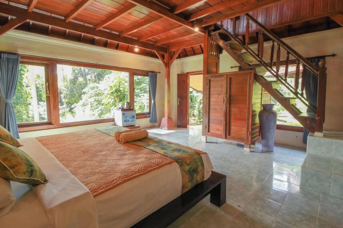 A spacious bedroom with a large bed, a wooden staircase, and a view of greenery outside.