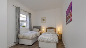 A bedroom with two beds, a window with curtains, a bedside table with a lamp, and a framed picture on the wall. - Seaside View Retreat in Clacton-on-sea (Clacton-on-Sea)
