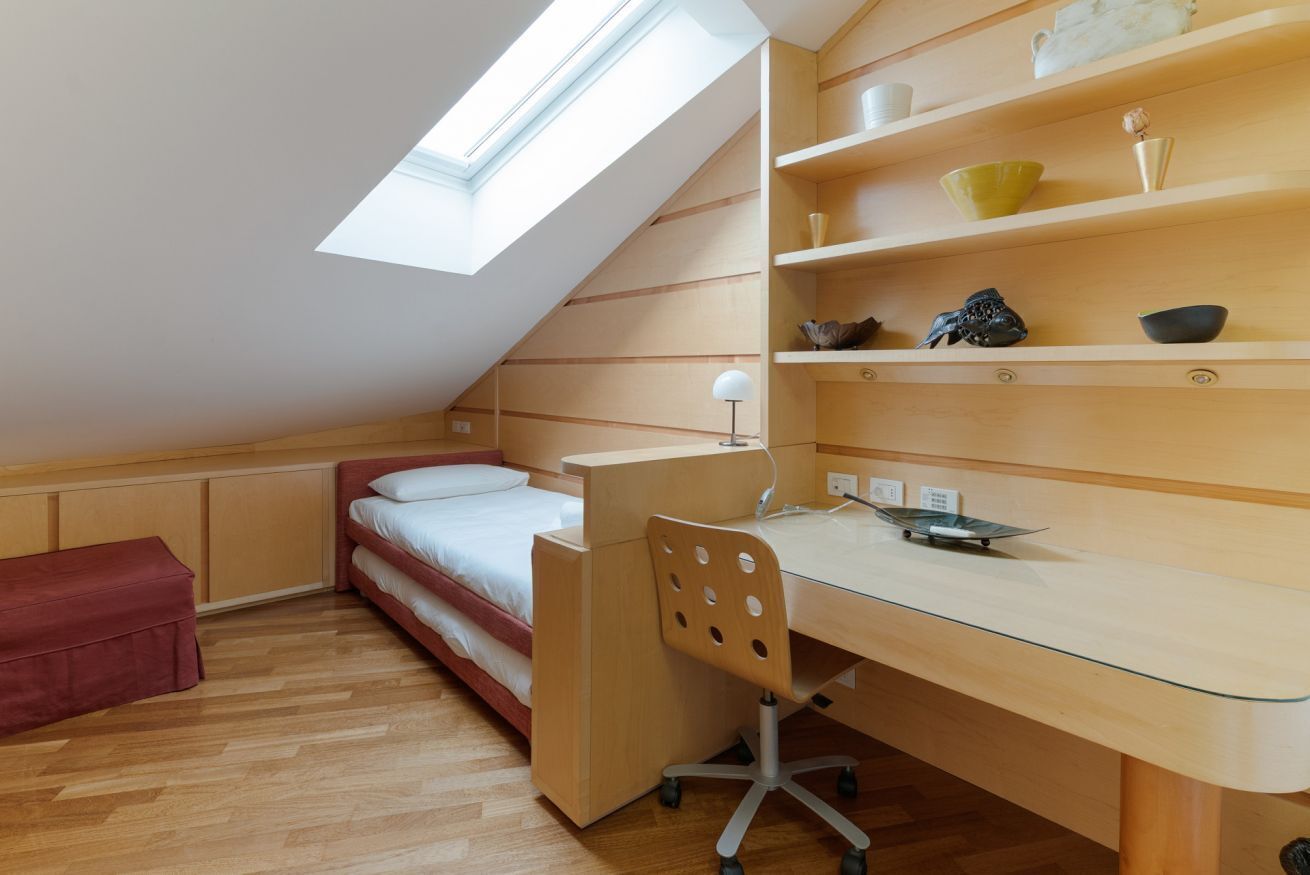 A compact room with a sloped ceiling, a bed, a desk with a chair, and shelves with various items.