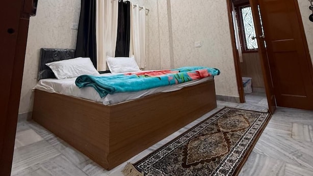 A bedroom with a bed, a patterned rug, a ceiling light fixture, and a door. - SHRI SHYAM KASHI STAY VARANASI (Varanasi)