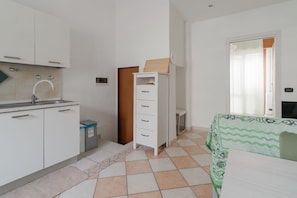 A small, clean room with a kitchenette, a bed with a patterned bedspread, and a window with curtains.