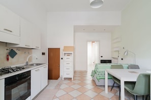 A compact kitchen with a stove, sink, and cabinets, a small dining area with a table and chairs, and a bed with a green patterned blanket. - GuestHost - A comfortable 40 square meters apartment, perfect for four people, located on the ground floor of a building.The property is located in a quiet area of ​​Milan and is well served by public transportation, allowing guests to easily explore (Milan)