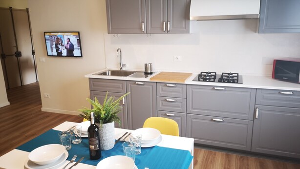 A modern kitchen with grey cabinets, a microwave, and a sink. There's a dining table set with plates, cutlery, and a bottle. A TV is mounted on the wall, and a plant is on the table. - Delicious next to Florence (Scandicci)