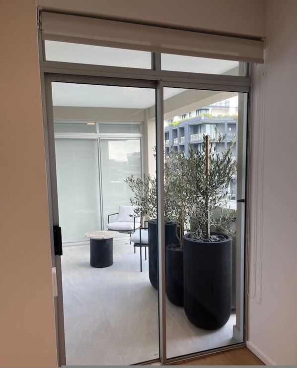 A modern interior with a sliding glass door leading to a balcony. The balcony has a minimalist design with a white chair and a round table. There are large potted plants on the balcony. - Casa Mia Lux - Sea Point 2 Bedroom Apartment (Cape Town)
