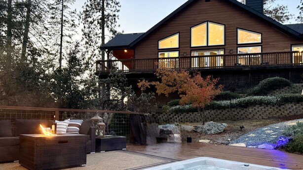 A hot tub with a wooden deck, outdoor furniture, and a large wooden building with multiple windows in the background. - Elegant Blackwood Grove by Casa Oso (Mariposa)