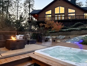 A hot tub with a wooden deck, outdoor furniture, and a large wooden building with multiple windows in the background. - Elegant Blackwood Grove by Casa Oso (Mariposa)