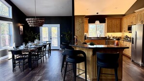 A modern kitchen with wooden cabinets, a central island, and a dining area with a large window. - Elegant Blackwood Grove by Casa Oso (Mariposa)
