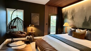 A bedroom with a large bed, a hanging chair, a small table with a lamp, and a painting of a forest on the wall. - Elegant Blackwood Grove by Casa Oso (Mariposa)