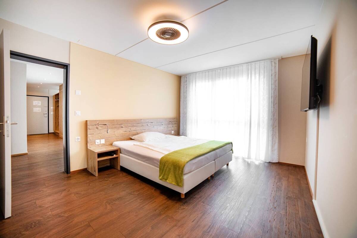 Habitació d'hotel amb terra de fusta, un llit amb llençol verd, una taula de nit, televisió penjada a la paret i una finestra gran amb cortines.