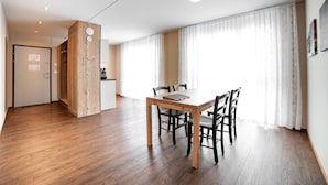 A modern dining area with a wooden table and chairs, a kitchen in the background, and large windows with sheer curtains. - Wohntel - wohnen wie im Hotel (Sevelen)