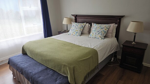 A neatly made bed with a green blanket, two matching pillows, and a wooden headboard. There are two bedside tables with lamps, and a window with curtains.