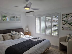 A bedroom with a bed, a ceiling fan, an air conditioning unit, a nightstand with a lamp, and a window with blinds. - Seaestaatmoffatquietbeachparklargefenced Yard (Sunshine Coast)
