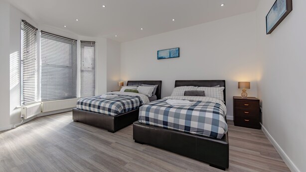 A bedroom with two beds, a radiator, a nightstand, and a wall-mounted picture. - Apartment - Sleeps 7 - Parking - Close to Pier (Clacton-on-Sea)