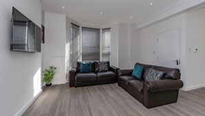 A modern living room with two dark brown leather sofas, a potted plant, and a large flat-screen TV mounted on the wall. - Apartment - Sleeps 7 - Parking - Close to Pier (Clacton-on-Sea)