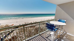 Een balkon met uitzicht op een strand, een tafel en blauwe stoelen.