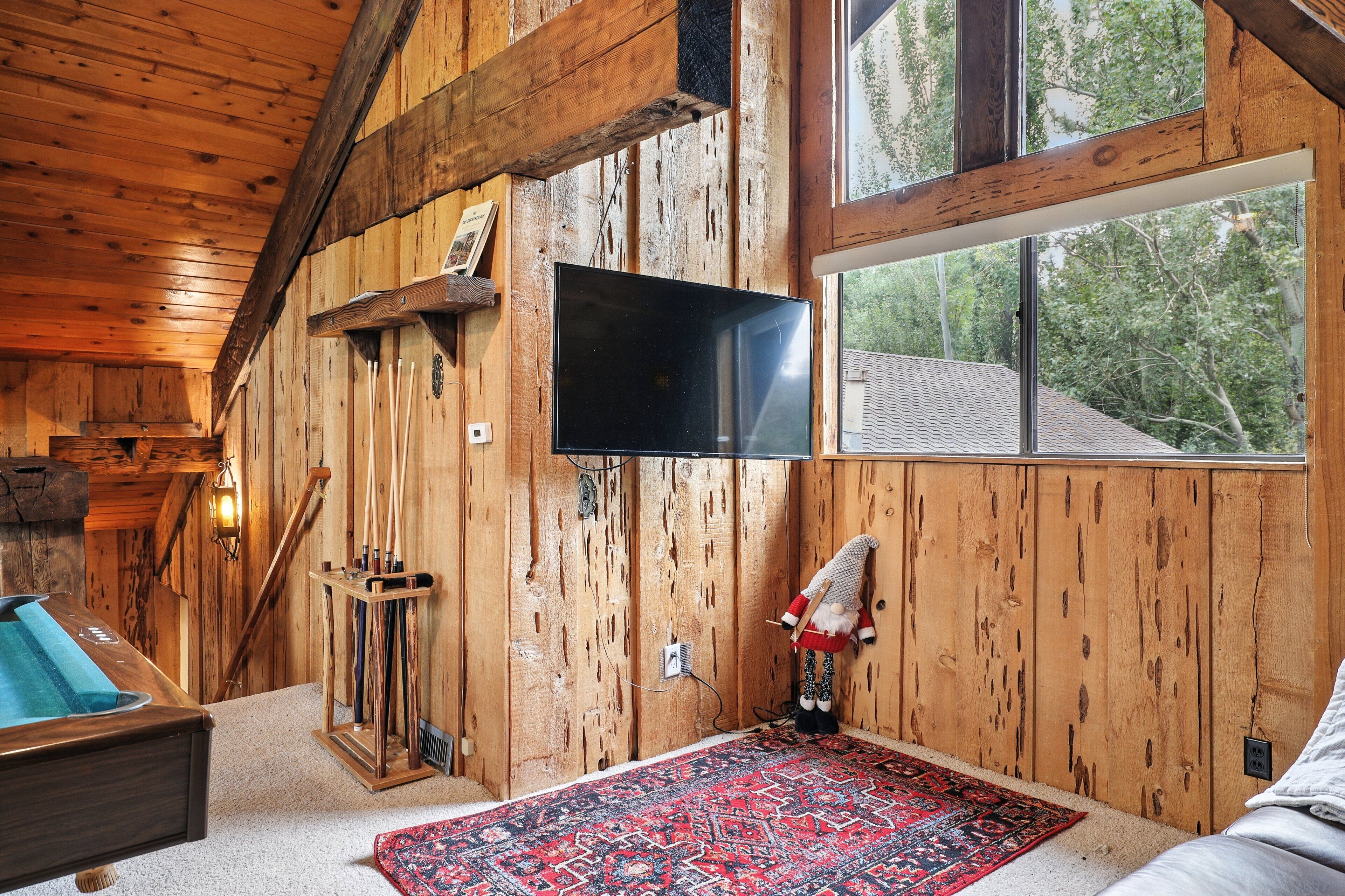 A wooden cabin interior with a television mounted on the wall, a pool table, and a large window offering a view of trees.