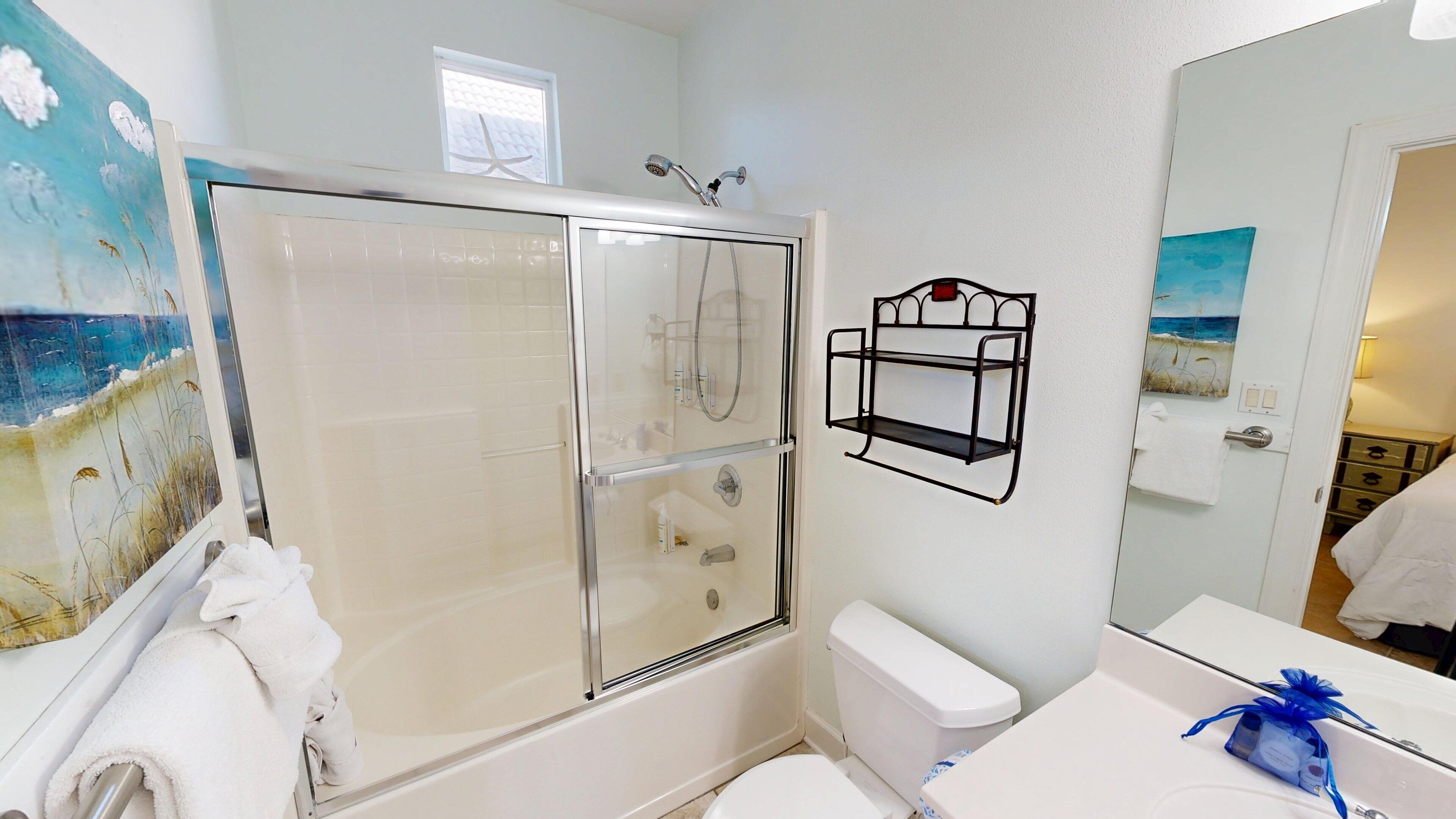 Une salle de bain avec une douche à parois de verre, des toilettes blanches, un lavabo avec une serviette bleue et un tableau sur le thème de la plage accroché au mur.
