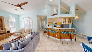 A spacious living area with a large sofa, a wooden coffee table, and a ceiling fan. There is a kitchen with a bar area featuring bar stools. - Mango Tango 5 Bedroom Home (Destin)