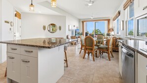 A spacious kitchen with a large island, dining area with wicker chairs, and a view of palm trees through large windows. - Big Island Na Hale O Keauhou C5 2 Bedroom Condo (Kailua-Kona)