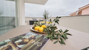 A table with a plate of lemons and olive branches, a glass of water, and a magazine on a balcony with a view of buildings. - Three-bedroom penthouse  Break with shared pool (Novalja)