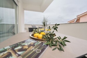Ein Tisch mit Zitronen, Olivenzweigen, einem Glas Wasser und einer Zeitschrift auf einem Balkon mit Blick auf Gebäude.