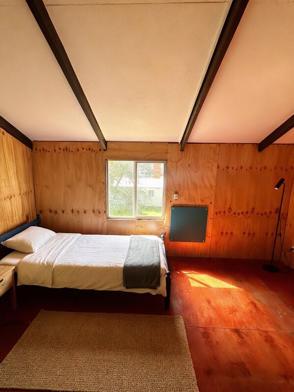 Une chambre avec un plancher en bois, un lit recouvert d’une courtepointe blanche, une fenêtre et un téléviseur fixé au mur.