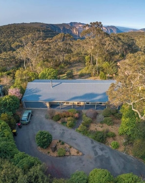 A large building with a flat roof, surrounded by dense vegetation and hills in the background. - Govetts Leap Lookout Cottage With Sauna (Blackheath)