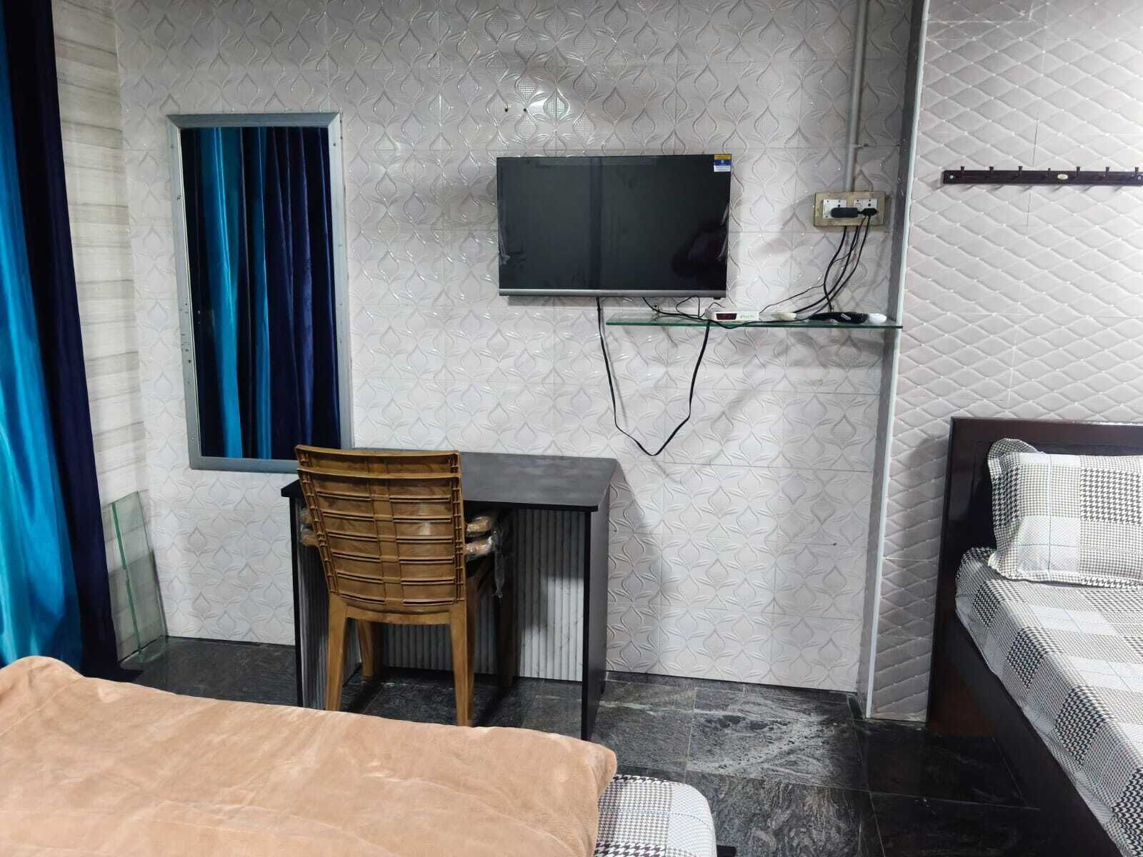 Una habitación con cama, escritorio y silla, televisión en la pared y ventana con cortinas.