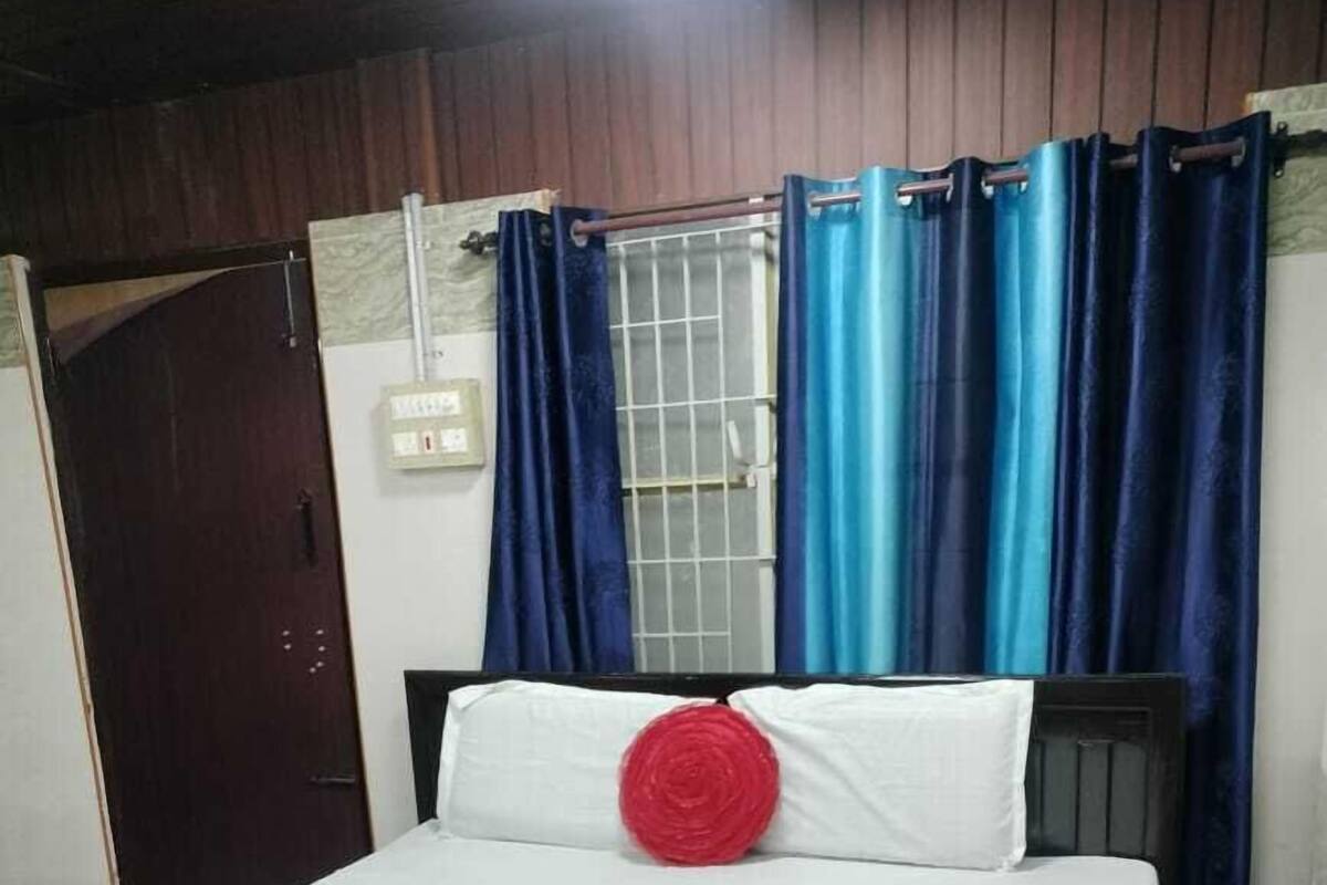 A bed with a white mattress, a red pillow, and a purple blanket. There is a wooden door and a window with blue curtains.