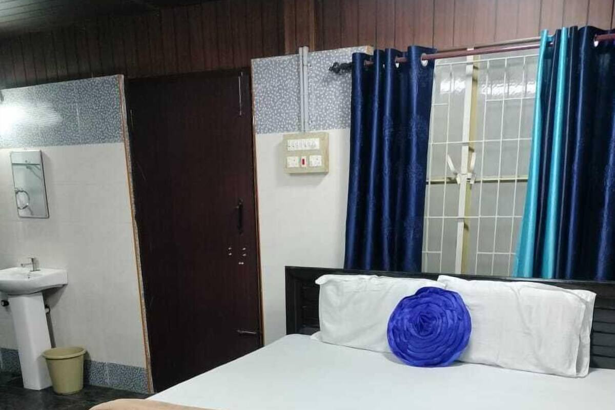 A bedroom with a bed, a bedside table, a window with curtains, a wall-mounted light switch, and a door.