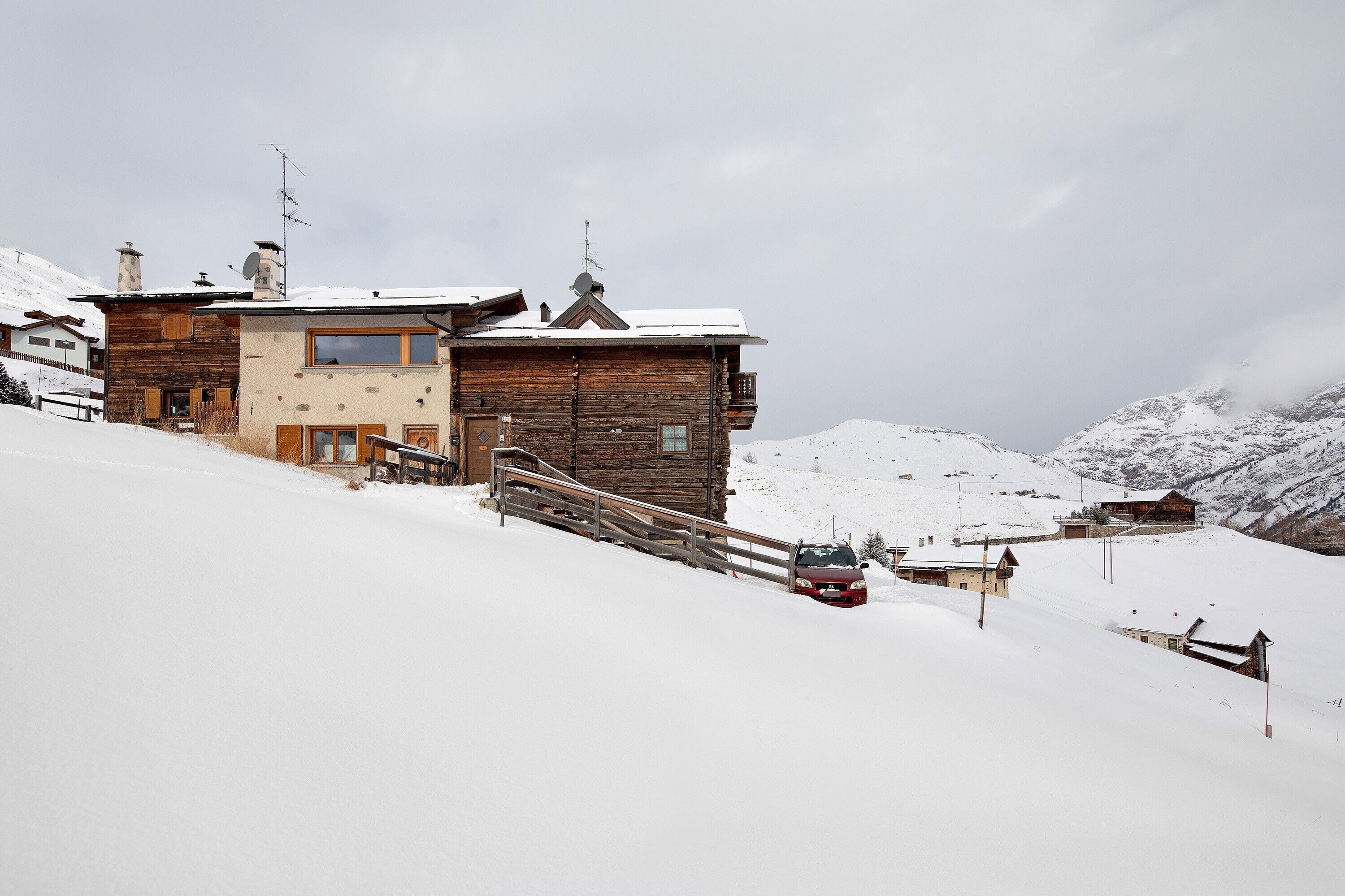 Un chalet en bois avec un escalier en bois, une voiture et une montagne enneigée en arrière-plan.