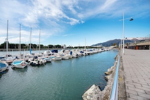Ein Yachthafen mit vertäuten Booten an der Uferpromenade, einem gepflasterten Gehweg und einem Berg im Hintergrund.