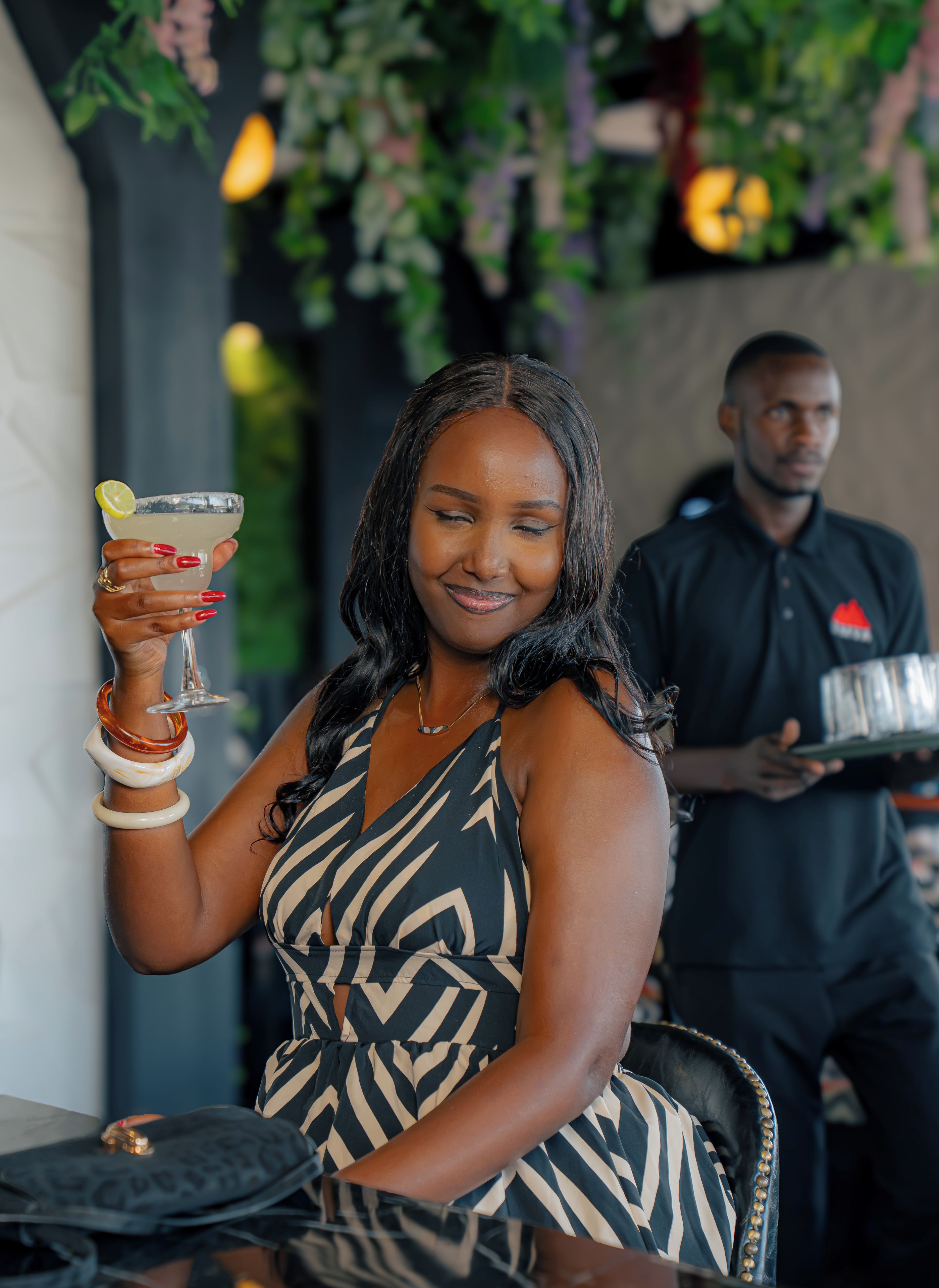 A person holding a drink with a lemon garnish, seated at a table with a chair that has a leather seat and a metal frame.