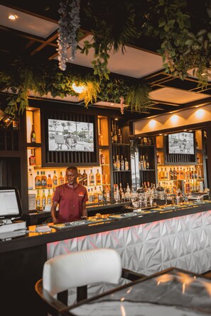 Zona de bar con un cantinero, estantes llenos de botellas de licor, una pantalla de televisión y plantas colgantes.