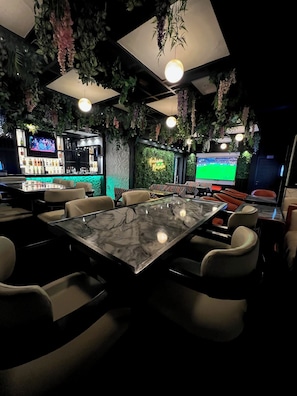 Un área de bar con asientos, una pantalla de televisión y plantas colgantes.