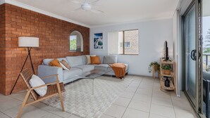 A modern living room with a brick accent wall, a grey sectional sofa, a wooden chair, a glass coffee table, and a floor lamp. - Layback on Marine (Kingscliff)