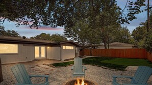 Ein Garten mit Feuerstelle, zwei Adirondack-Stühlen, einem Haus mit Garage und einem Holzzaun.