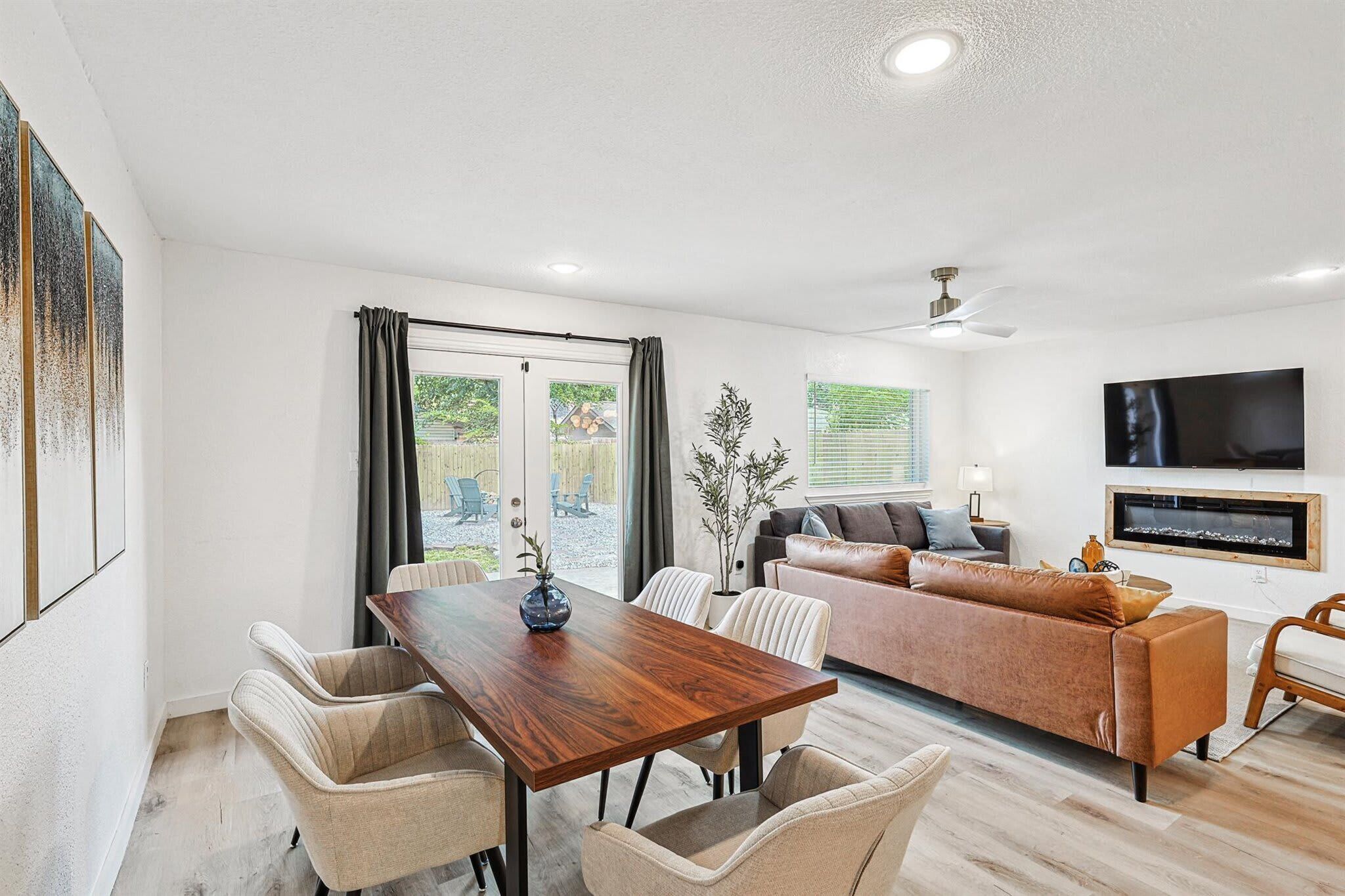 A modern living room with a wooden dining table, beige upholstered chairs, a brown sofa, and a flat-screen TV mounted on the wall.