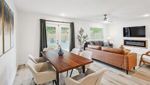 Ein modernes Wohnzimmer mit einem Holzespeisetisch, beigefarbenen Polsterstühlen, einem braunen Sofa und einem wandmontierten Flachbildfernseher.