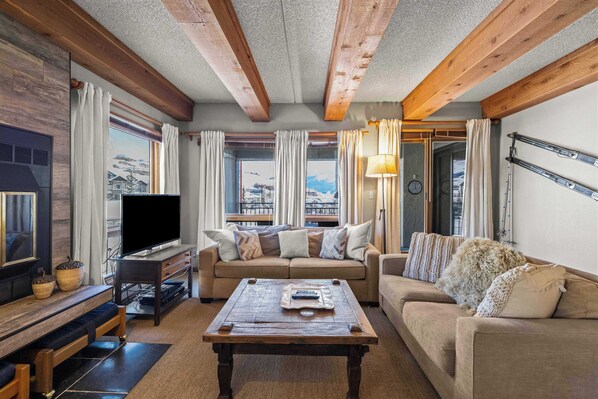 A cozy living room with a sofa, a coffee table, and a TV. There are windows with curtains, a fireplace, and a view of a snowy landscape. - The Plaza 231 | Walk to CBMR Lifts | 2BR Sleeps 6 (Crested Butte)